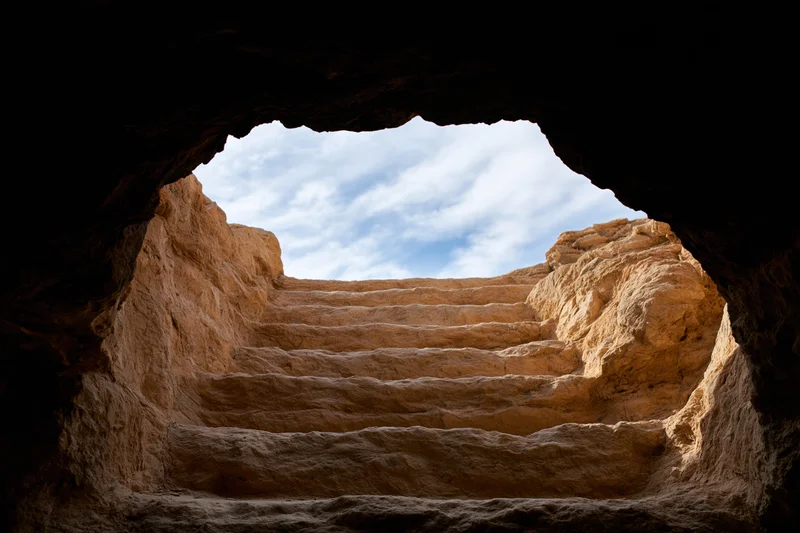 View from inside a dark stone cave looking out through an opening at a carved stone staircase leading up to a sky with clouds