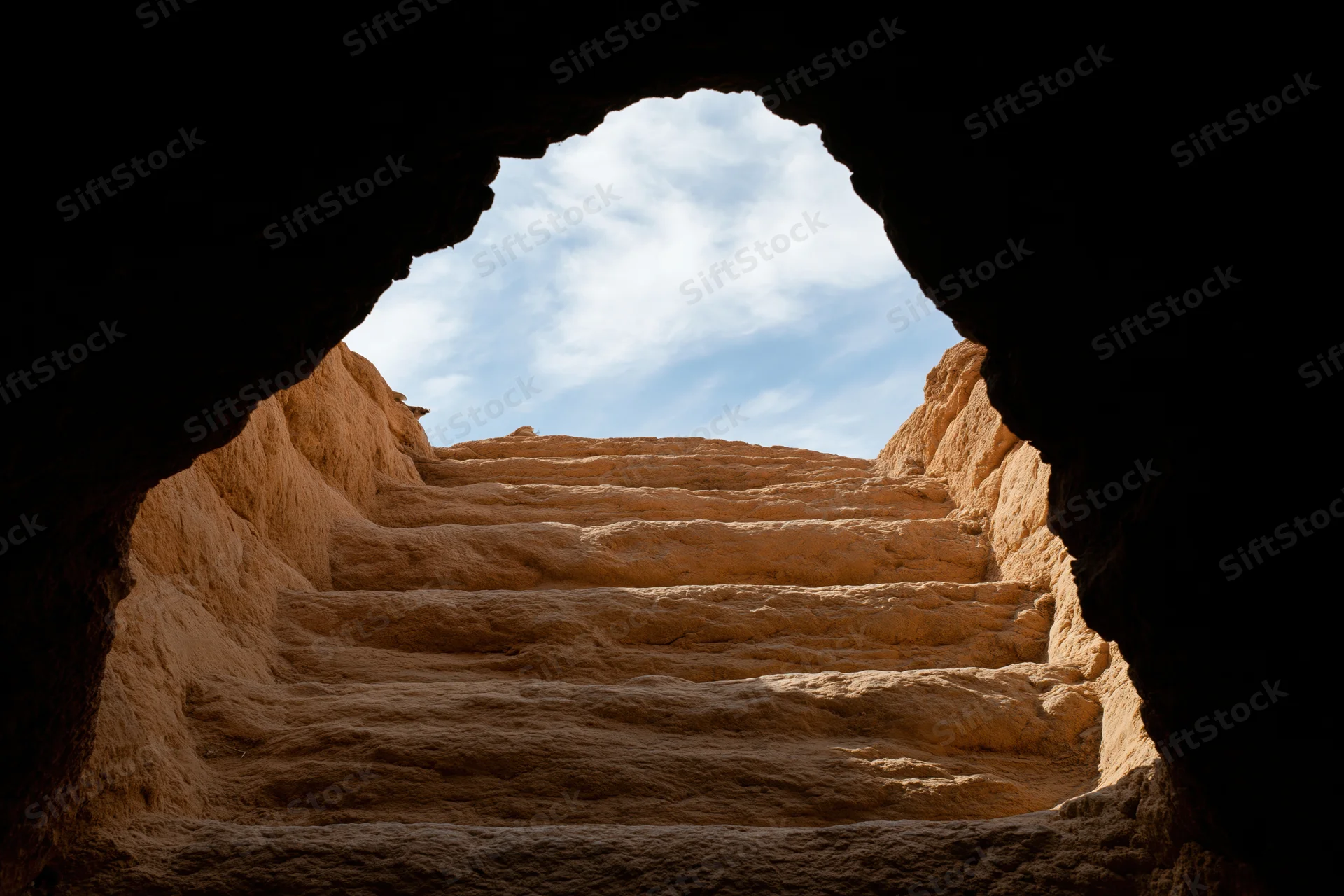View from inside a dark stone cave looking out through an opening at a sunlit sandstone staircase under a blue sky with clouds
