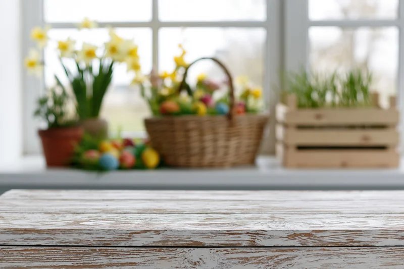 Weathered white wooden table surface in sharp focus with blurred easter decorations and spring flowers on windowsill in background
