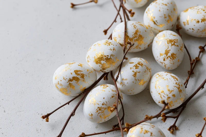 White easter eggs with gold leaf splatter markings arranged on branches on a light surface