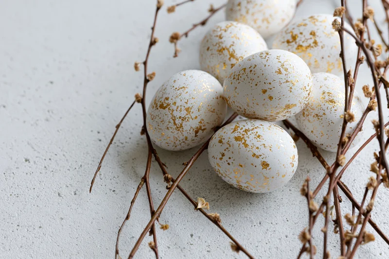 White easter eggs with gold leaf splatter markings arranged on concrete surface with dried branches and buds