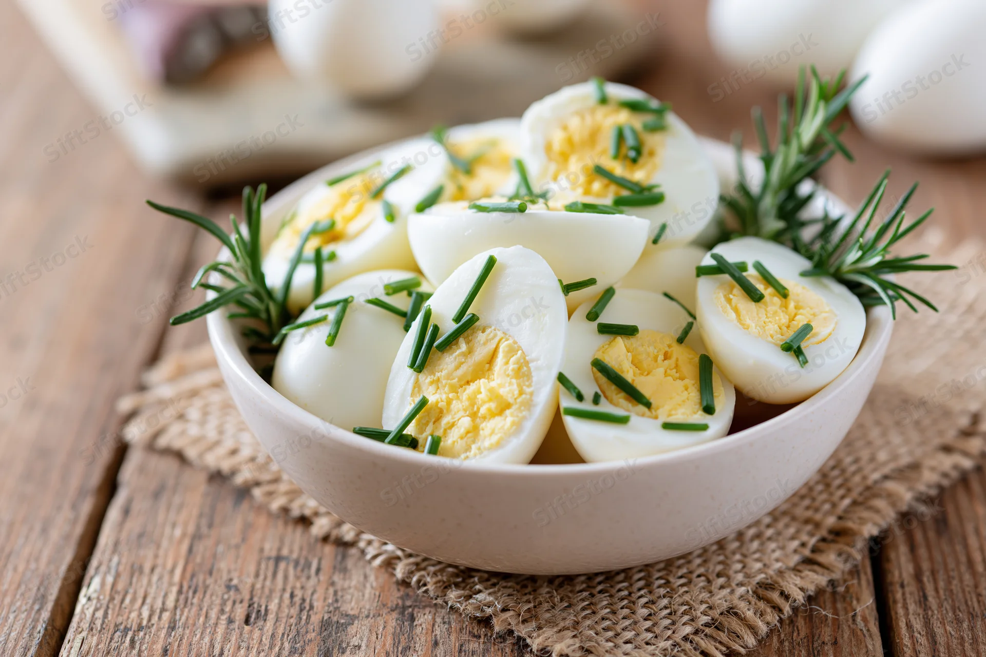 Halved hard-boiled eggs garnished with chives and rosemary in a white ceramic bowl on a wooden table with burlap placemat