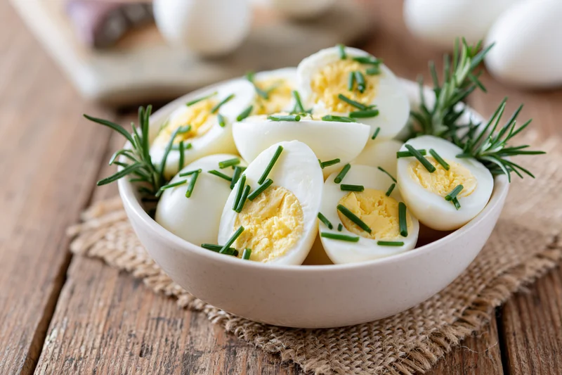 Halved hard-boiled eggs garnished with chives and rosemary in a white ceramic bowl on a wooden table with burlap placemat