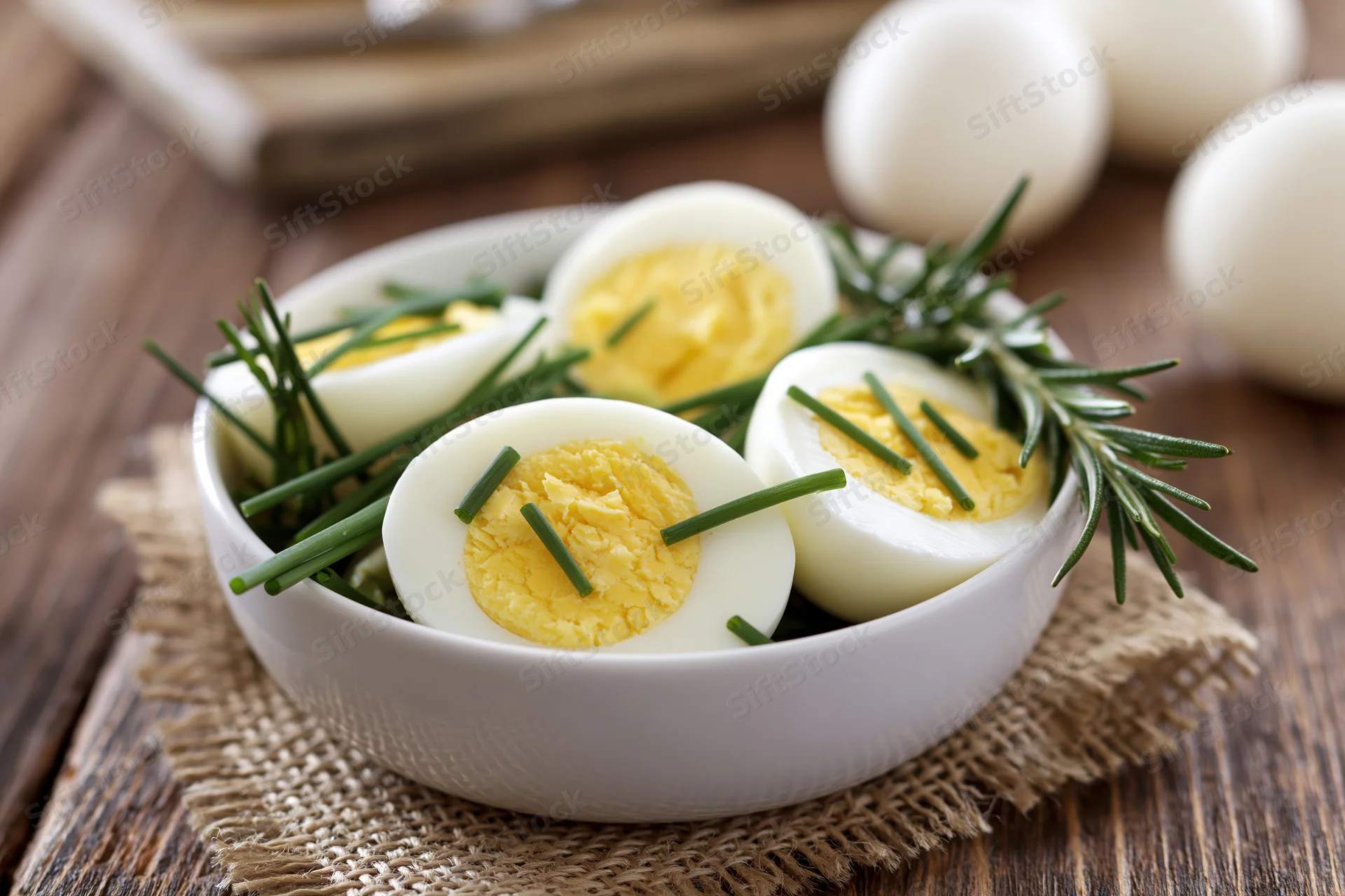 Halved hard-boiled eggs garnished with chives and rosemary in a white ceramic bowl on a wooden table
