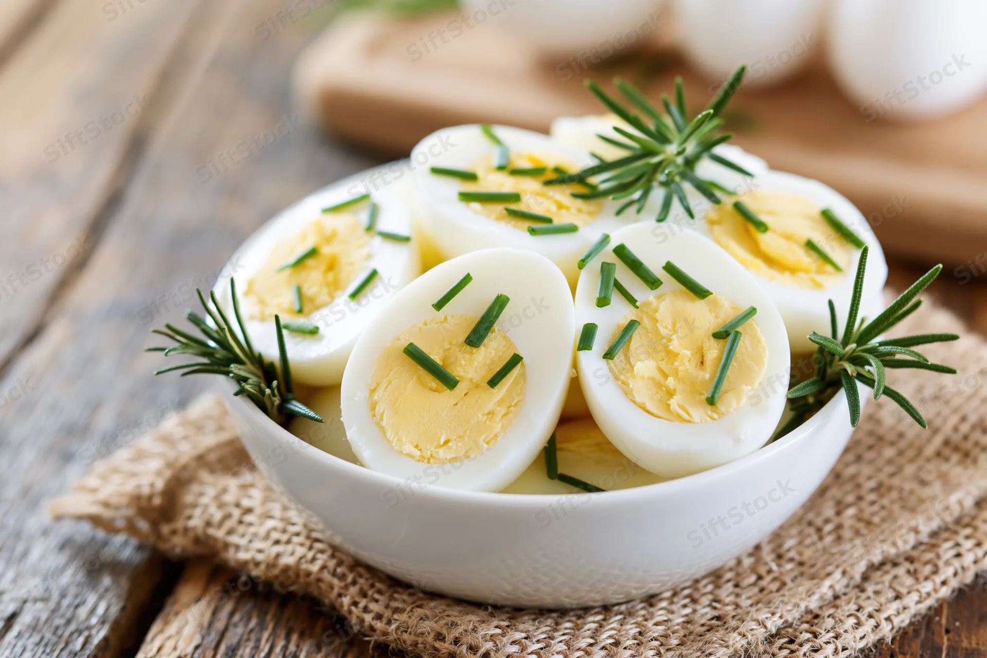 Halved hard-boiled eggs garnished with rosemary and chives in a white ceramic bowl on a rustic wooden table with burlap placemat