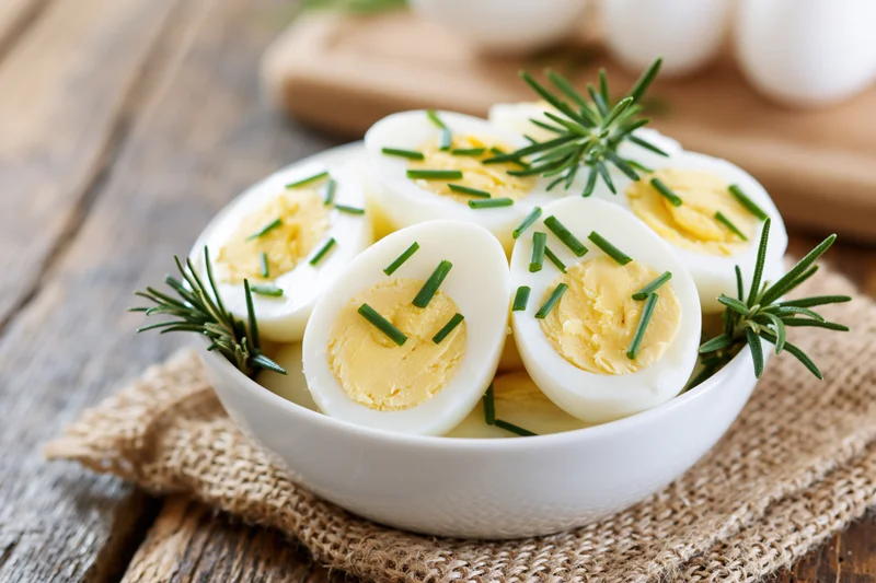 Halved hard-boiled eggs garnished with rosemary and chives in a white ceramic bowl on a rustic wooden table with burlap placemat