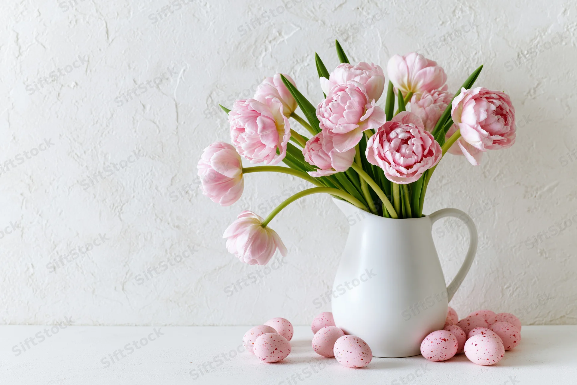 Pink double tulips arranged in a white ceramic pitcher on a table with speckled easter eggs around it
