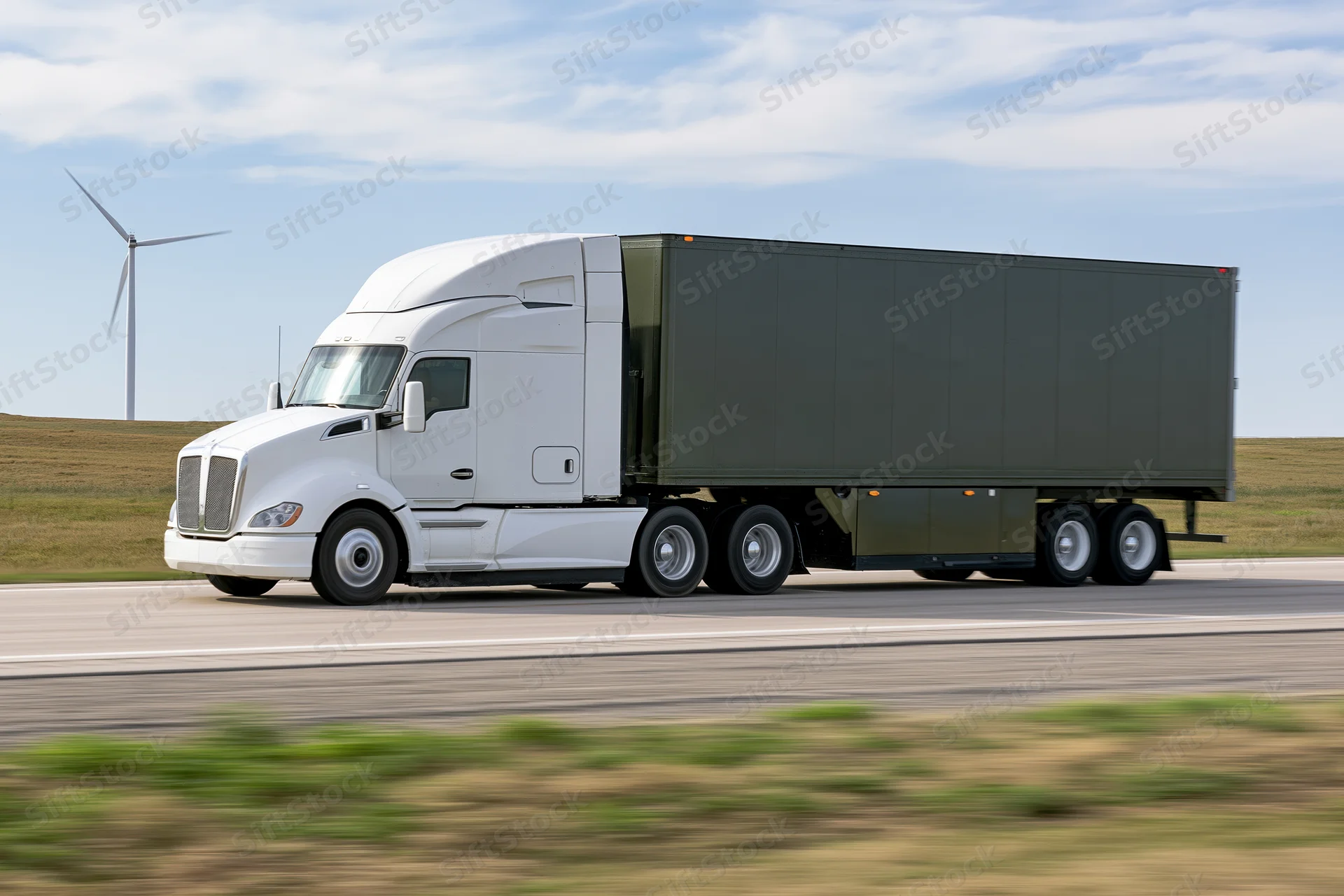White electric semi-truck hauling dark green trailer on highway with wind turbine in rural landscape