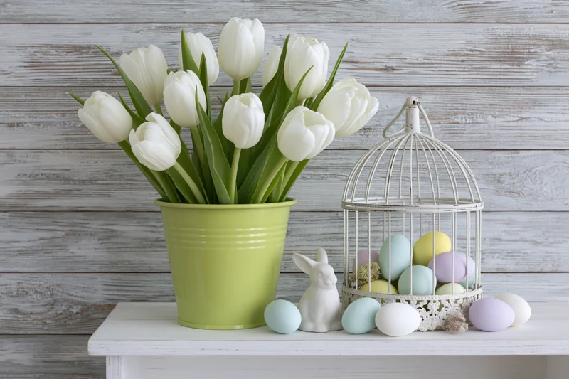 White tulips in a lime green bucket vase arranged on a white table with easter eggs and a ceramic rabbit beside a birdcage