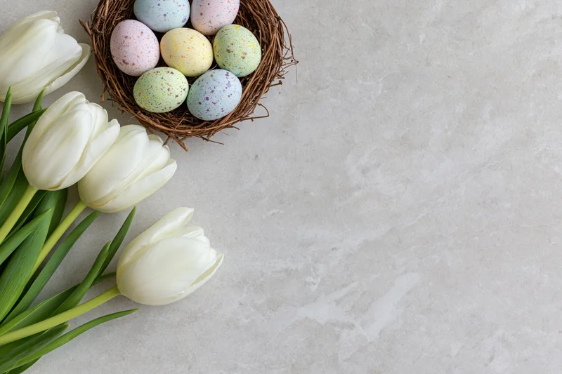 White tulips with green stems arranged beside a nest of speckled pastel eggs on a light stone surface