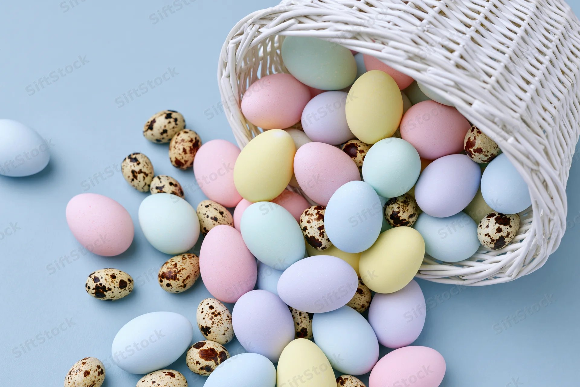 White wicker basket tipped over spilling pastel-dyed eggs and quail eggs onto a light blue surface