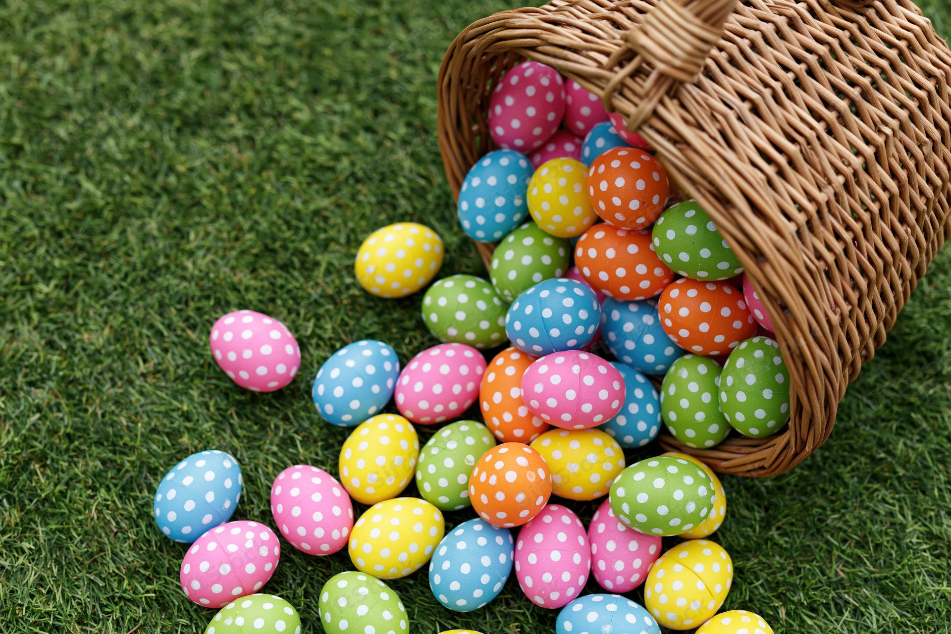 Wicker basket tipped over spilling colorful polka-dotted easter eggs onto green grass