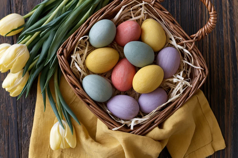 Wicker basket filled with hand-painted pastel easter eggs nestled in straw beside yellow tulips on wooden table
