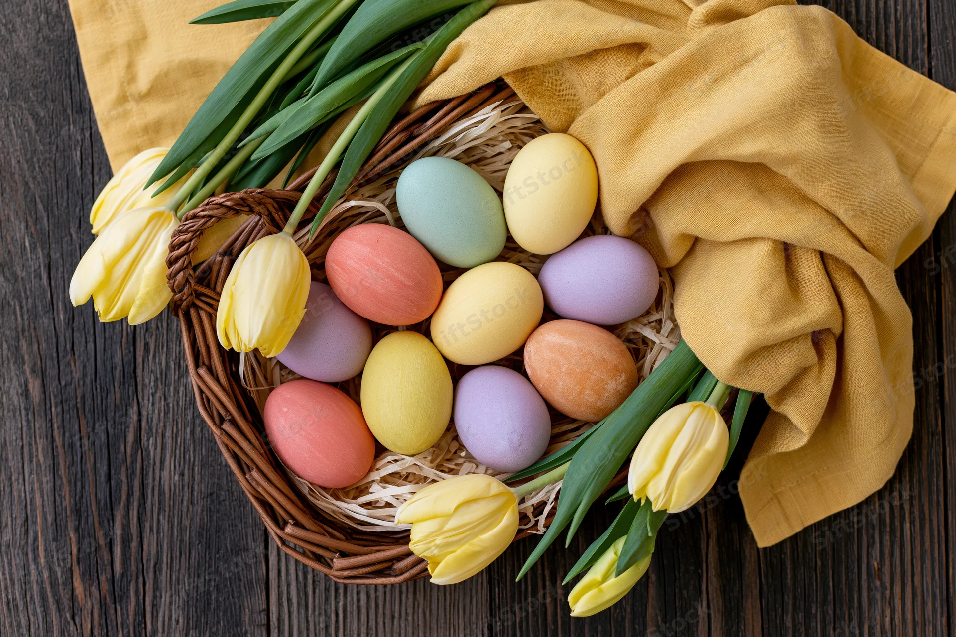 Wicker basket filled with hand-painted pastel easter eggs and yellow tulips on dark wooden table with mustard cloth