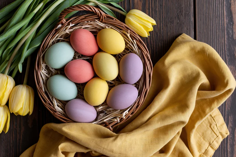 Wicker basket filled with hand-painted pastel easter eggs on wooden table with yellow tulips and cloth napkin
