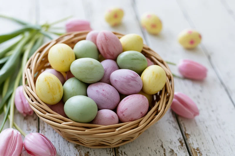 Wicker basket filled with pastel-painted easter eggs and pink tulips on a rustic wooden table