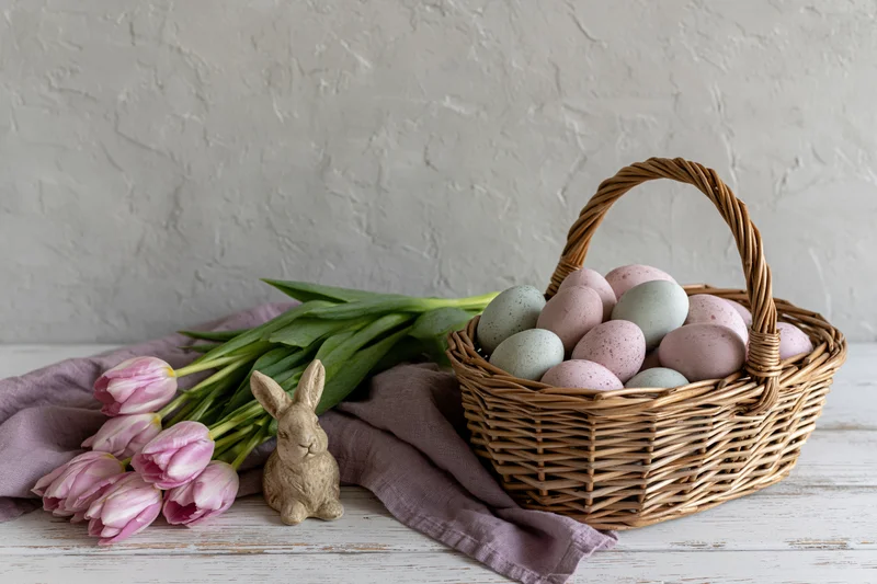 Wicker basket filled with pastel easter eggs on a white wooden table beside pink tulips and a bunny figurine