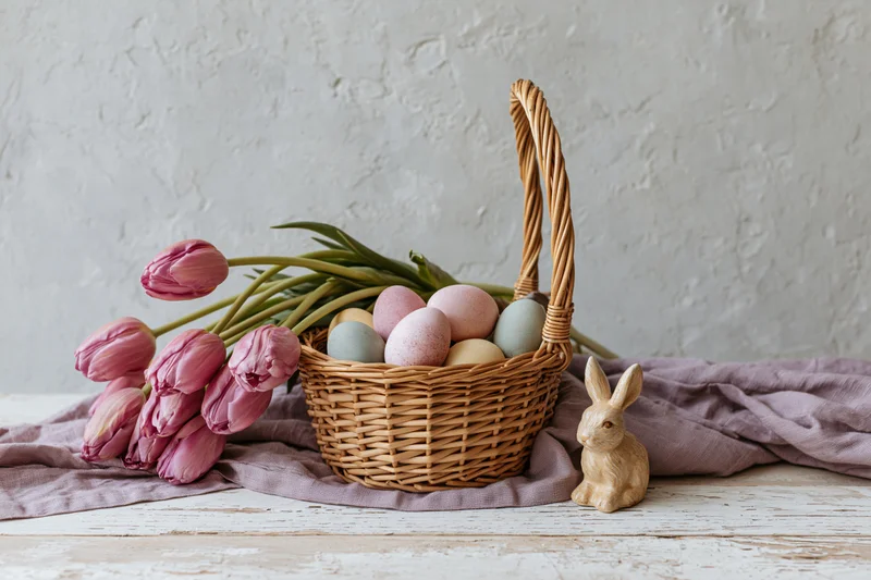 Wicker basket filled with pastel easter eggs and pink tulips on a cloth-covered table with a wooden bunny beside it