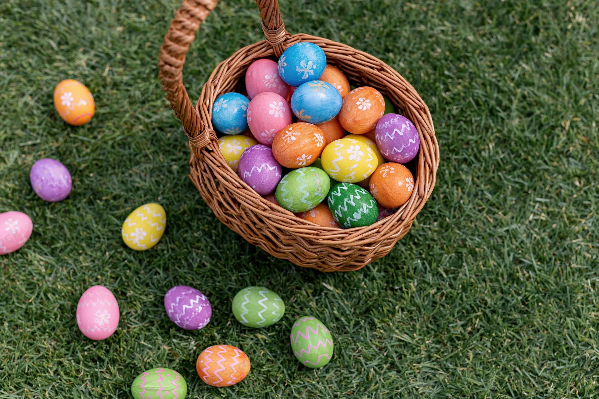 Wicker basket overflowing with hand-painted easter eggs scattered on green grass