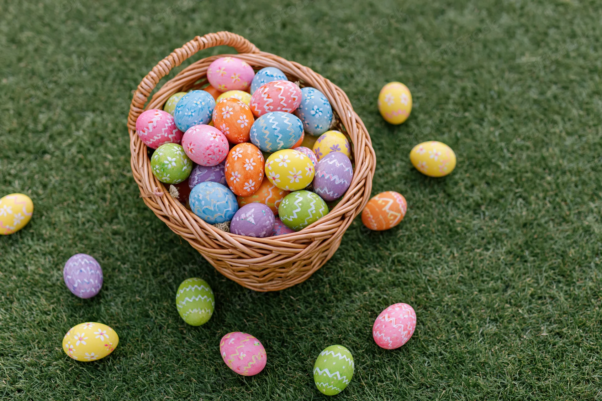 Wicker basket overflowing with hand-painted easter eggs scattered on green grass