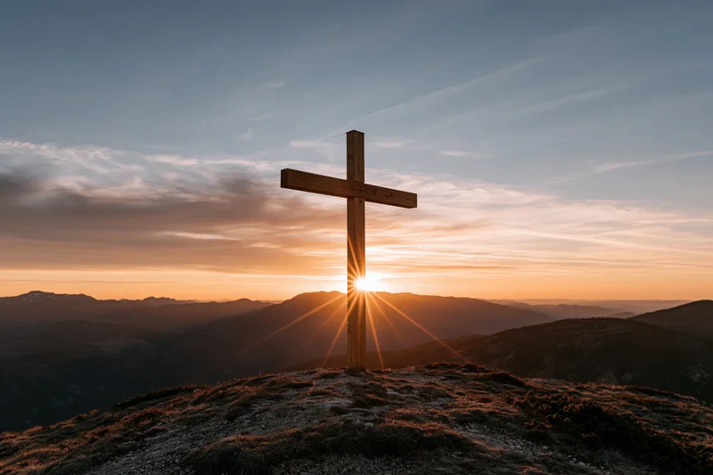 Wooden cross silhouetted on a hilltop at sunset with sun rays behind it over mountainous landscape