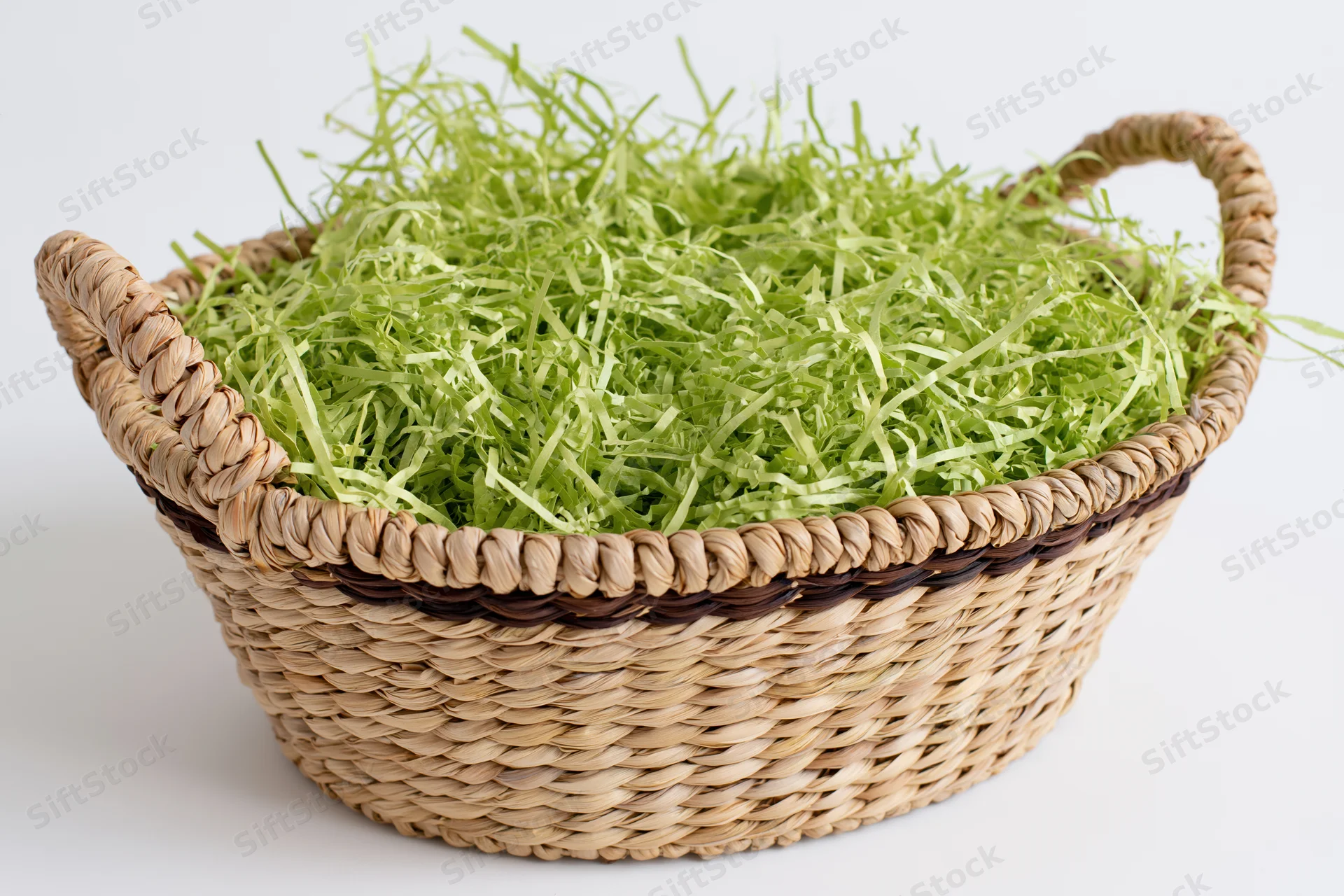 Woven wicker basket with two handles filled with green shredded grass on white background