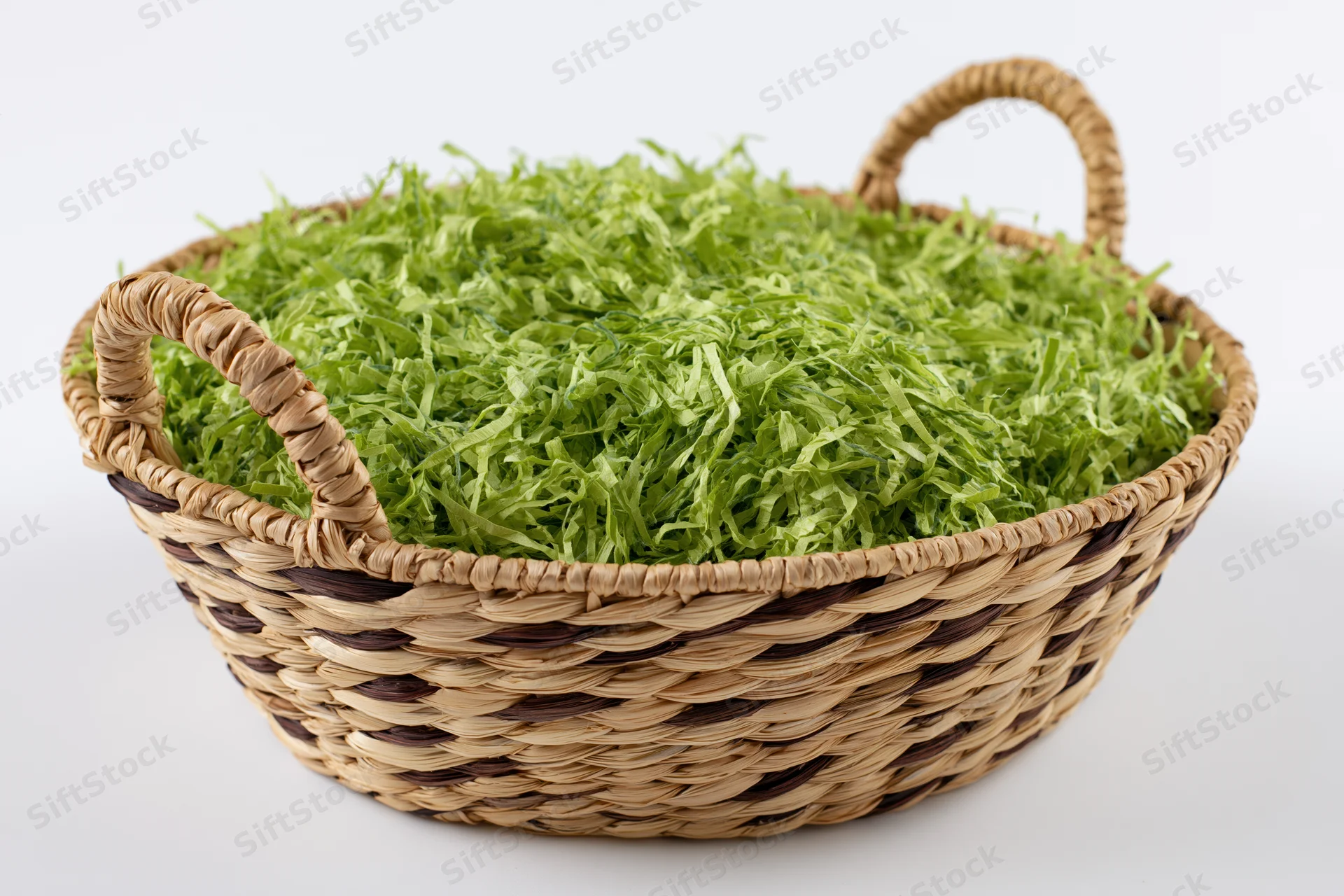 Woven wicker basket with two handles filled with bright green shredded paper on white background