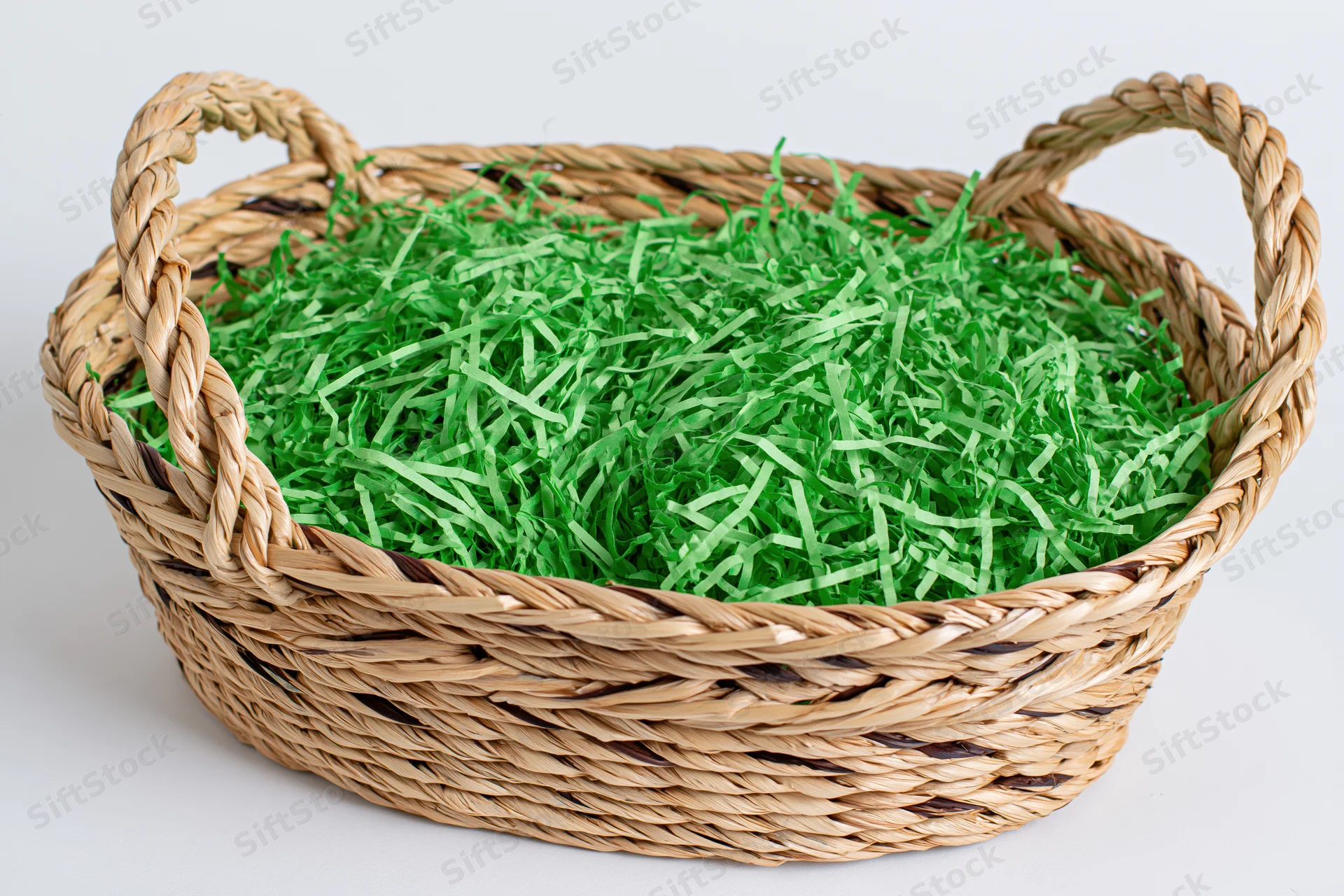 Woven wicker basket with two handles filled with bright green shredded paper filler on white background
