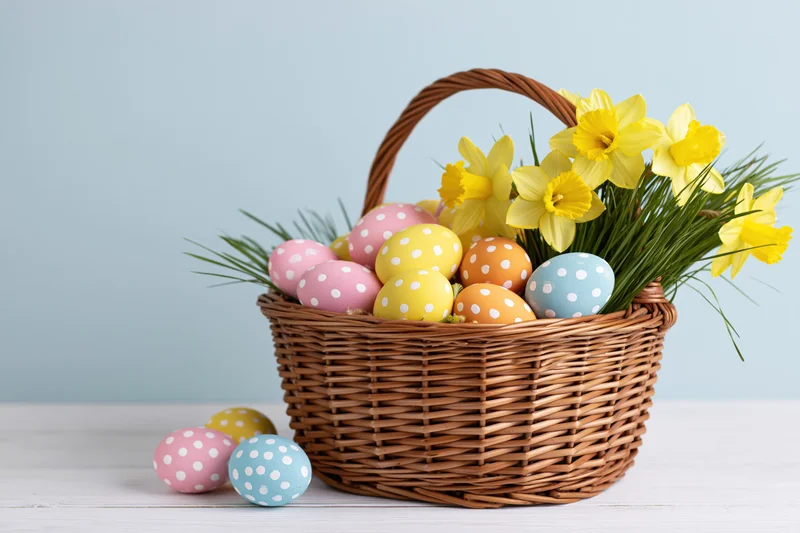 Wicker basket filled with pastel polka-dotted easter eggs and yellow daffodils on a white surface