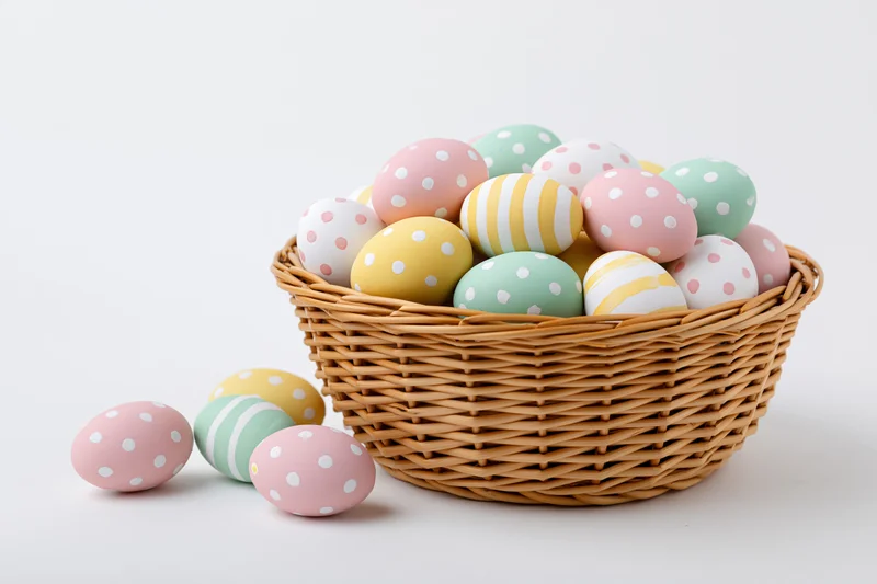 Wicker basket filled with pastel-painted easter eggs arranged on white surface with few scattered beside it