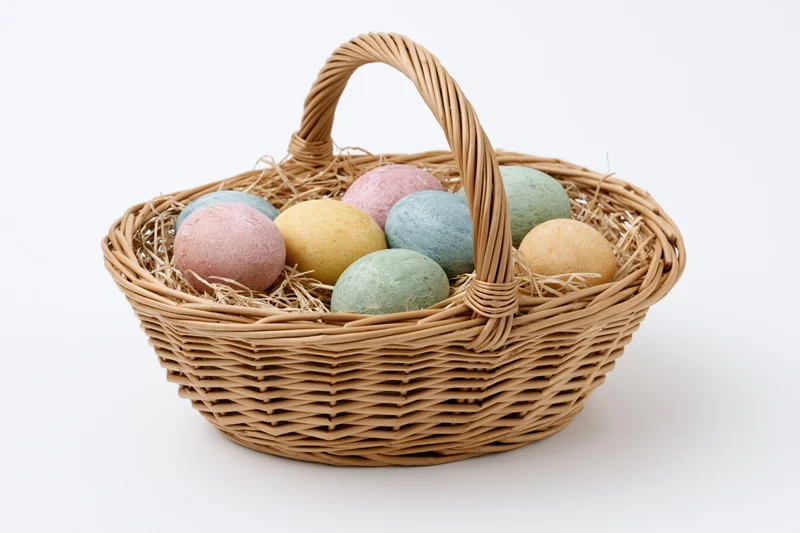 Woven wicker basket with looped handle holding pastel-colored eggs nestled in straw on white background