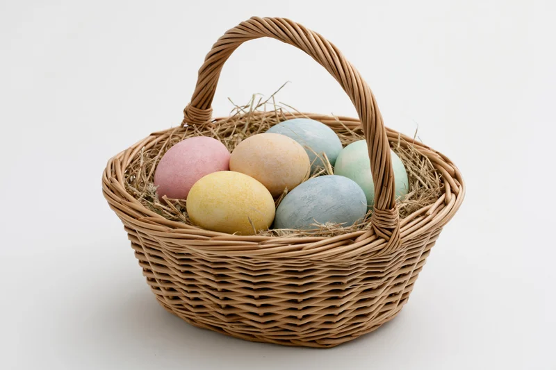 Woven wicker basket with looped handle holding pastel-colored eggs nestled in straw on white background