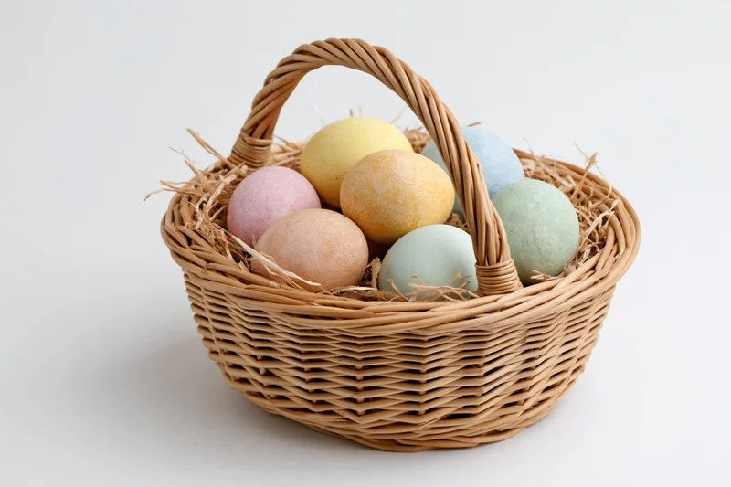 Woven wicker basket with looped handle filled with pastel-colored eggs on straw bedding against white background