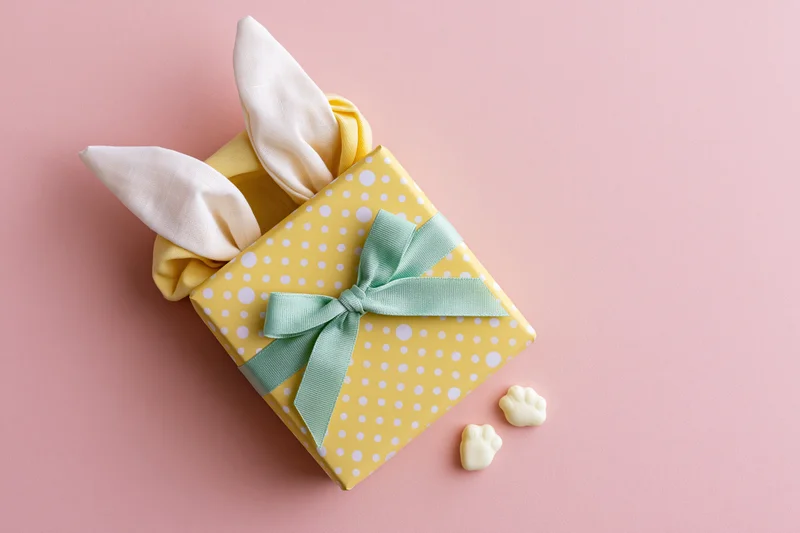 Yellow polka-dot gift box with white fabric bunny ears and mint green ribbon on pink background