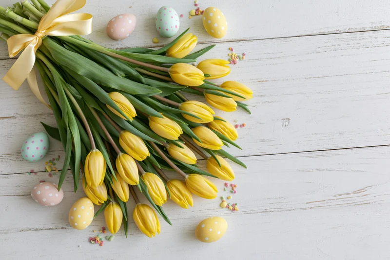 Yellow tulip bouquet tied with satin ribbon resting on white wooden table with decorated easter eggs and confetti scattered around