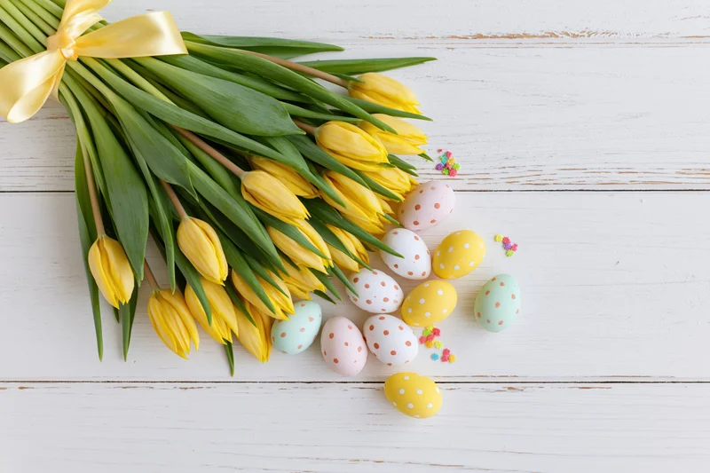 Yellow tulip bouquet tied with satin ribbon resting on white wooden surface beside decorated easter eggs