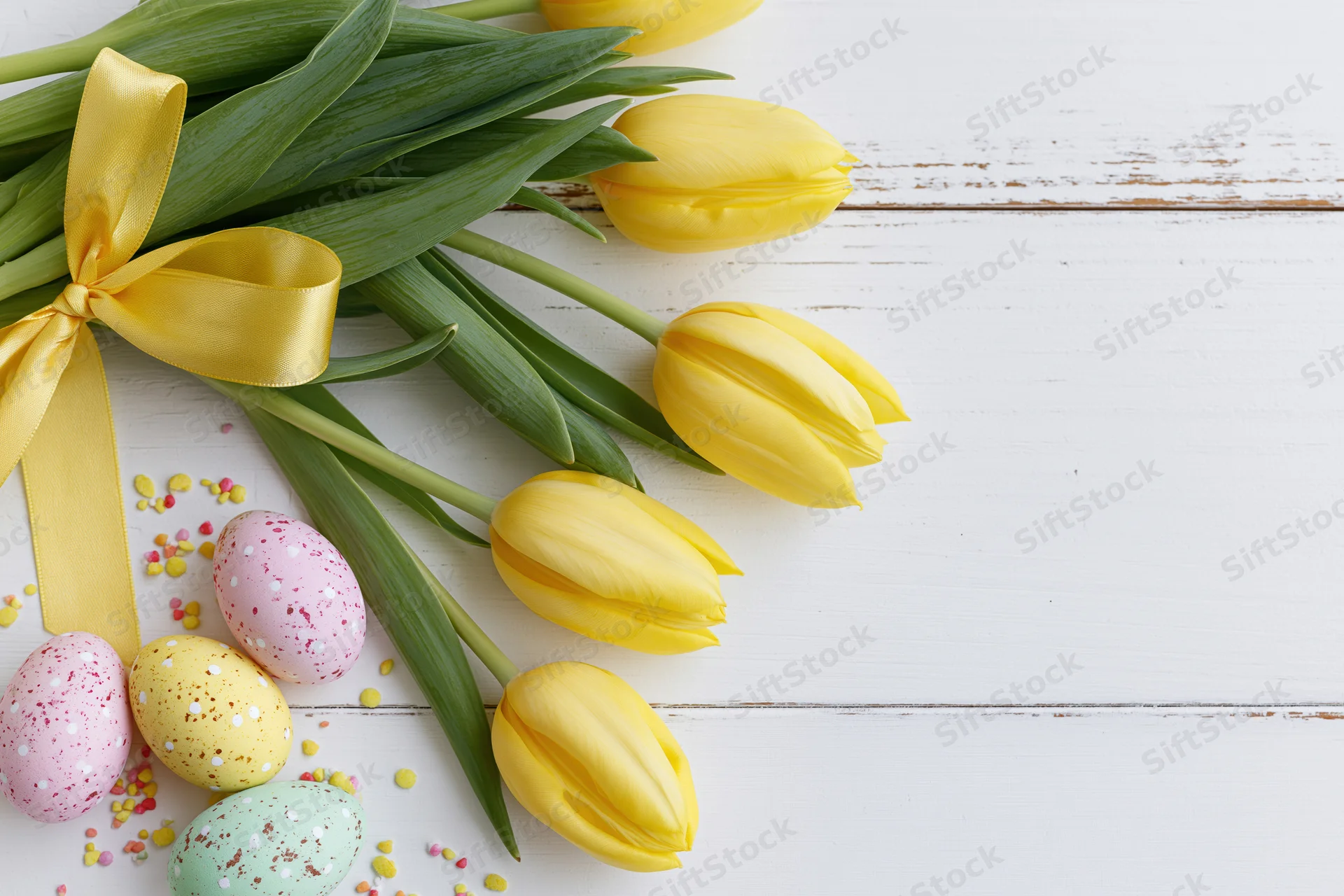 Yellow tulips with green stems tied with a satin yellow ribbon arranged with decorated easter eggs on a white wooden surface