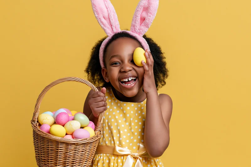 Young black girl with afro wearing pink plush bunny ears, laughing while holding a wicker basket of colorful easter eggs and playfully covering one eye with a yellow egg against a yellow background