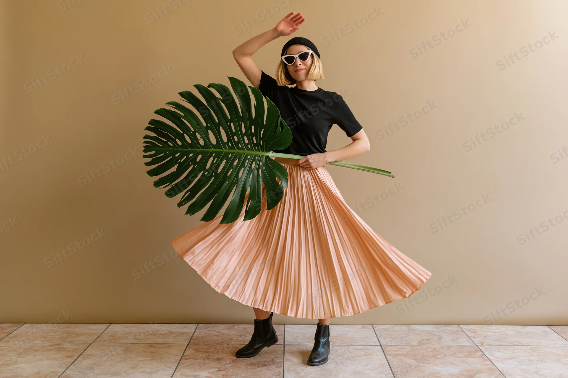 Young blonde woman dancing and twirling in a peach pleated skirt holding a large monstera leaf indoors