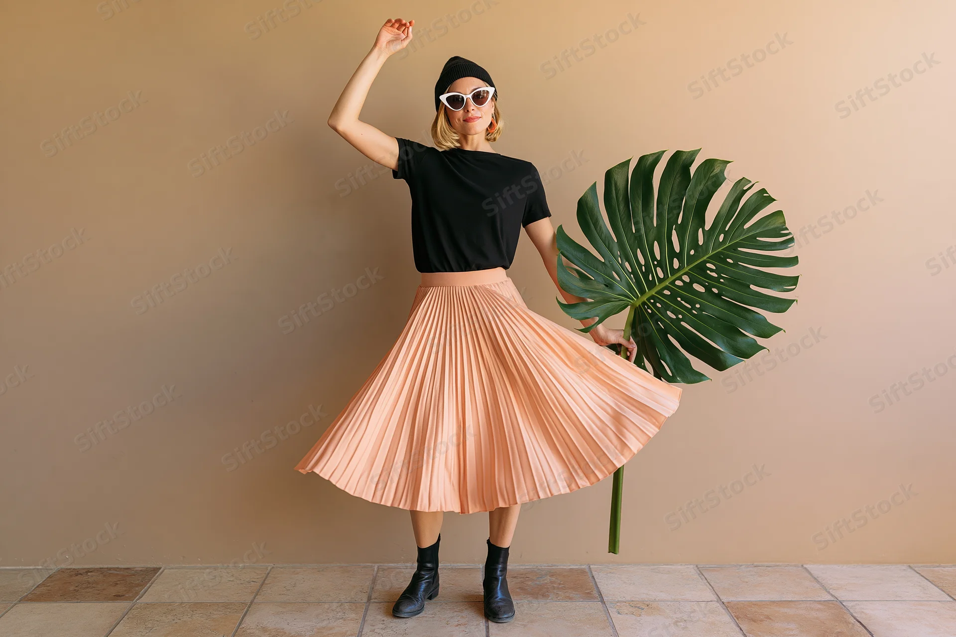 Young blonde woman dancing and twirling in a pleated peach skirt while holding a large monstera leaf indoors