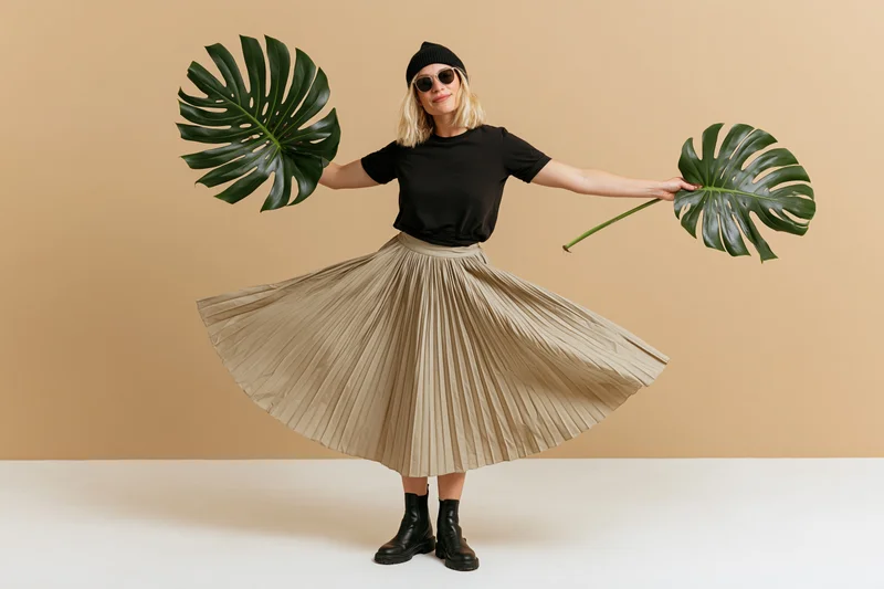Young blonde woman spinning in pleated skirt holding monstera leaves in studio