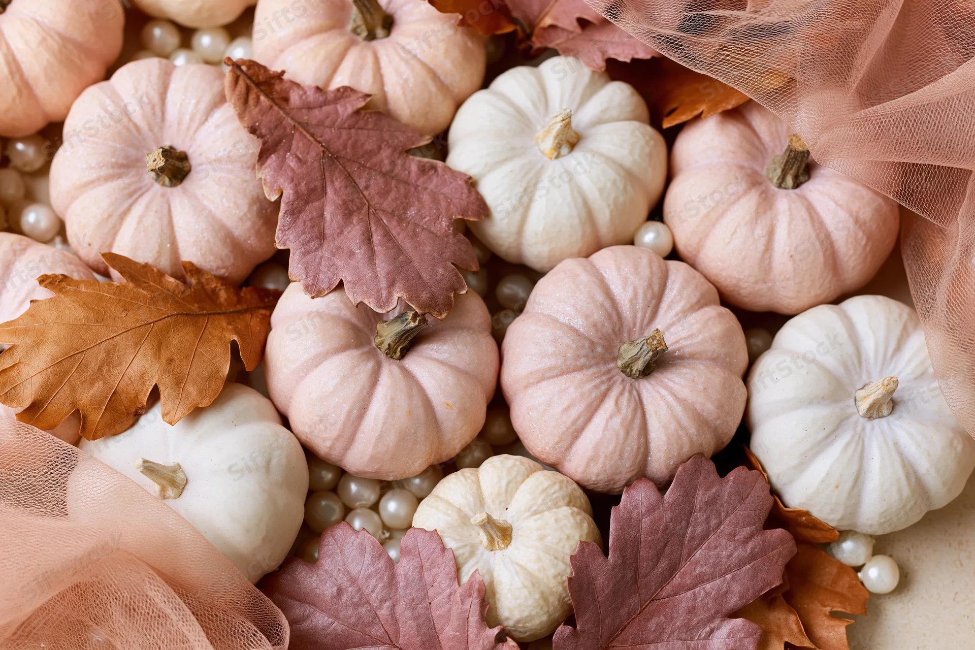 Small decorative pumpkins and autumn leaves arranged in a flat lay with pearls and tulle fabric on a light surface