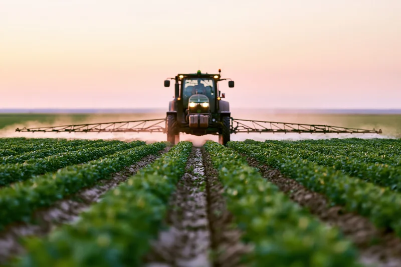 Tractor with boom sprayer moving through rows of crops in a field at dusk