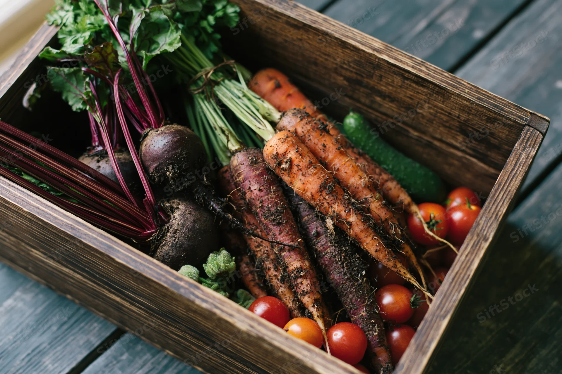 Wooden crate filled with freshly harvested vegetables including carrots, beets, tomatoes, and cucumbers on a wooden surface