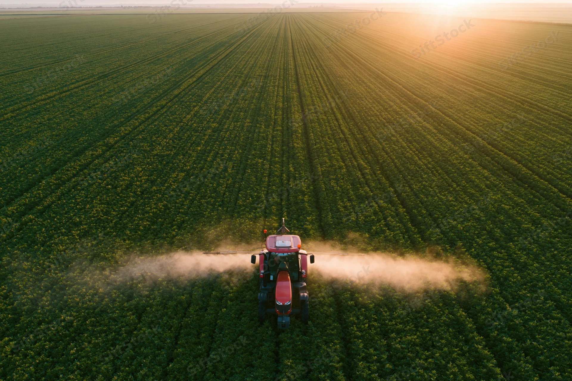 Red tractor spraying pesticide mist across a vast green crop field from aerial view at sunset
