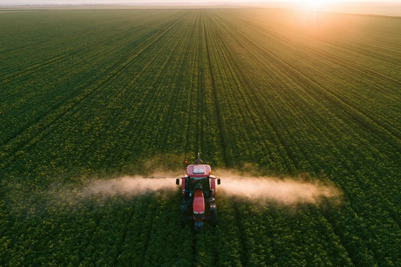 Red tractor spraying pesticide mist across a vast green crop field from aerial view at sunset
