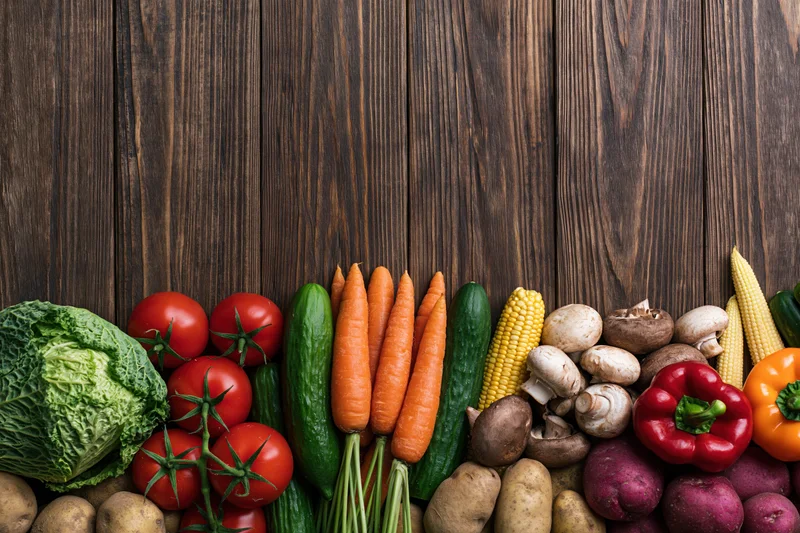 Assorted fresh vegetables arranged in a dense row along the bottom of a dark wooden surface with copyspace above