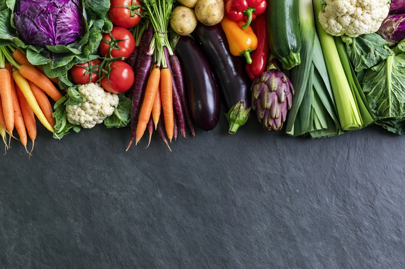 Assorted fresh vegetables arranged in a border along the top of a dark slate surface with copyspace below