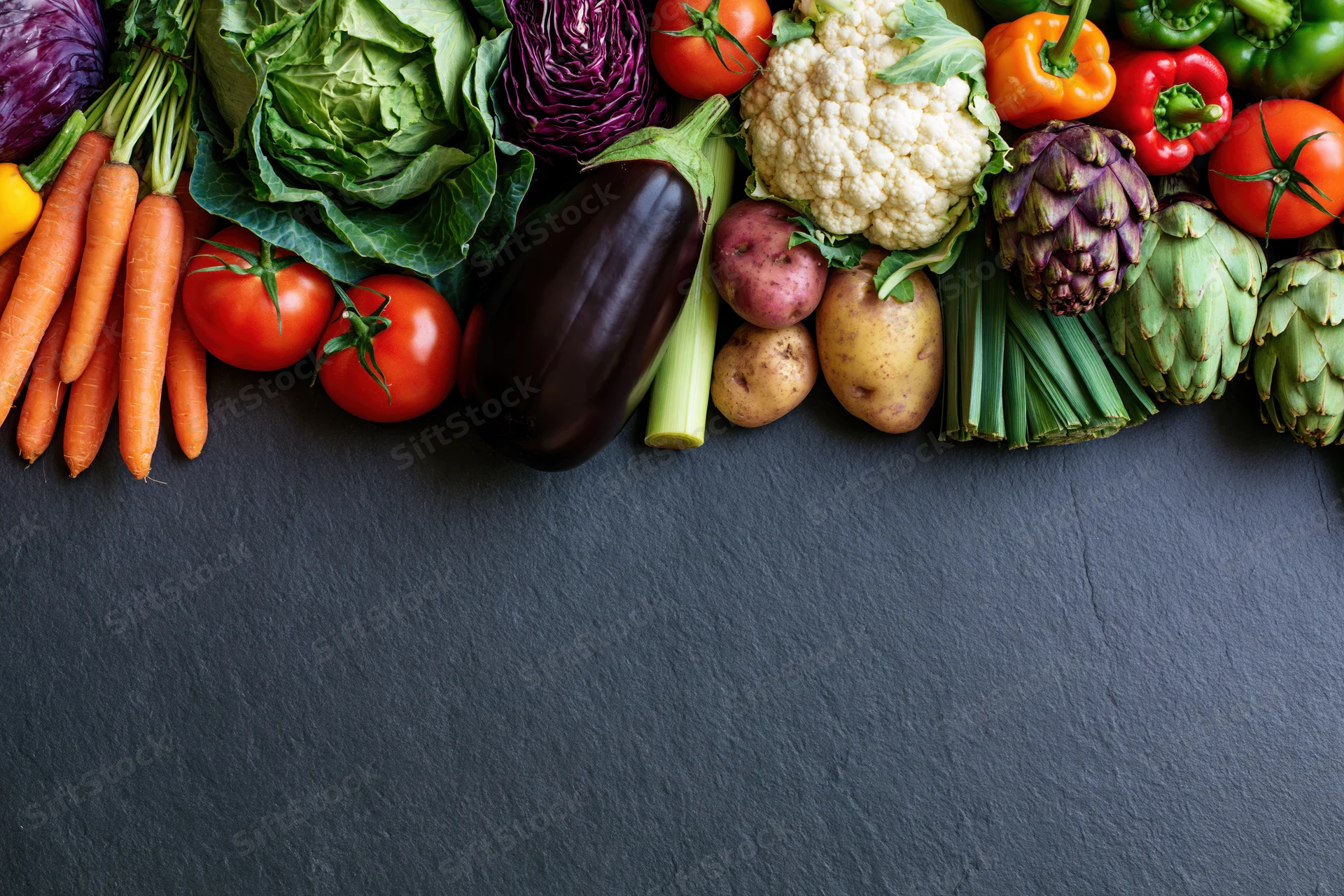 Assorted fresh vegetables arranged in a border along the top of a dark slate surface with copyspace below