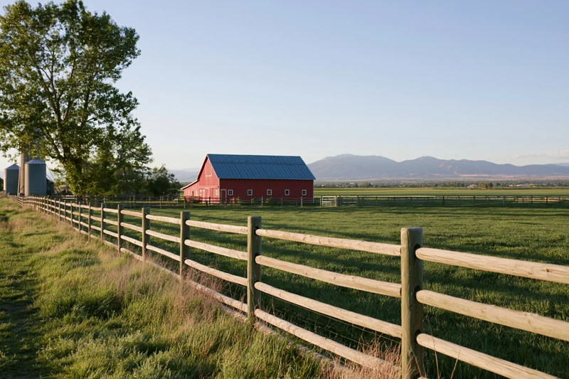 Red barn with blue metal roof in a grassy field bordered by wooden fence with mountains in the distance